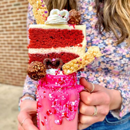 A pink milkshake in a Mason jar with a piece of red cake, icing and sweet treats piled on top of the rim