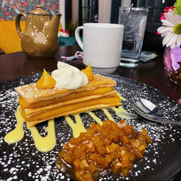Photo of a table with a pitcher and cup of coffee, flowers and cup of water, and in the foreground, their orange Pumpkin Napoleon, with kabocha purée, pastry, spiced pumpkin compote, all sprinkled with powdered sugar