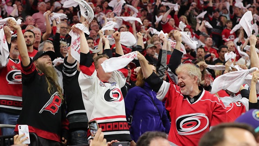 A large crowd of hockey fans standing and waving towels in the air, most are wearing Carolina Hurricanes jerseys, cheering as if a goal has just been scored