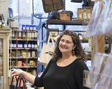 Pam Blondin, a white woman dressed in a black blouse, smiles at the camera between the aisles of her store DECO Raleigh. Gift items like purses, candles, etc. surround her.