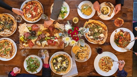 A top-down view of a wooden dining table filled with a dozen plates of food, with several diners reaching their arms in for a slice of pizza or sampling of other appetizers
