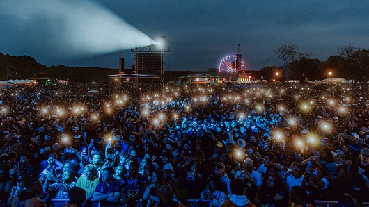 Large crowd facing stage fills open field at Dorothea Dix Park at night, with a blue glow cast across faces and a Ferris wheel lit up in the far background