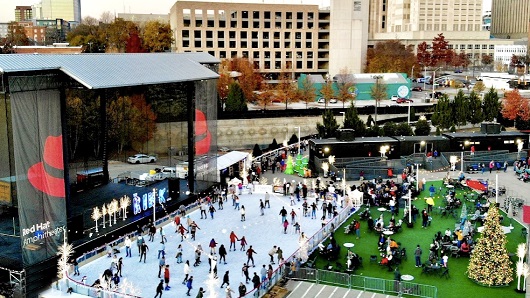 A drone photo of THE RINK at Red Hat Amphitheater, a large ice skating rink in downtown Raleigh filled with 50 or so skaters and more people hanging out by firepits and heaters outside of the rink