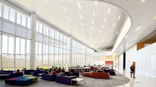 A large, open area at Hunt Library, with two-story glass windows lining the wall on the left letting in natural light, and colorful and unique chairs spread throughout the 3,000-square-foot space. One person on the camera's right is walking towards viewer 