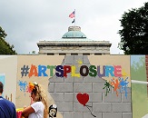 A Banksy-style mural is on large posterboard is displayed in front of the North Carolina State Capitol, with #artsplosure written in various colors across the top and a red, heart-shaped balloon depicted floating against a gray brick wall