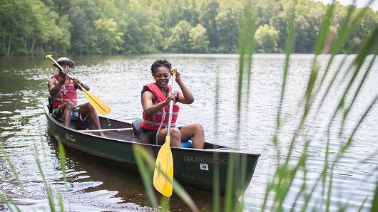 Woman and man wearing life vests paddle a canoe on a calm lake with tall grass in the foreground