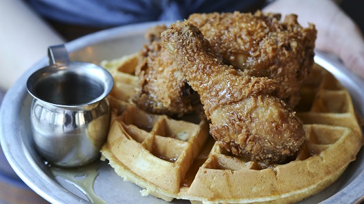 A metal plate is held out to the camera, displaying a circular, Belgian-style waffle with fried chicken sitting atop and a small cup of syrup off to the side