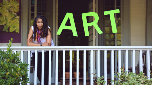 Lilnda Dallas, a black woman with long hair wearing a white top with no sleeves, looks at camera from a front porch with a neon green sign that reads 'ART' to the right of her