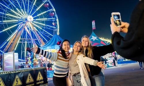 Three women posing for a picture in front of a lit-up Ferris wheel at night at the fair