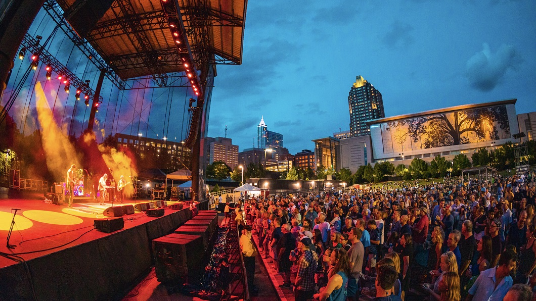 View from side of stage at Red Hat Amphitheater, with large crowd standing and watching band and view of Raleigh skyline at dusk in the background