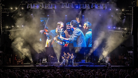 A bluegrass band jams on stage in front of a huge crowd at Red Hat Amphitheater, well after the sun has set, with purple and blue lights glowing on stage