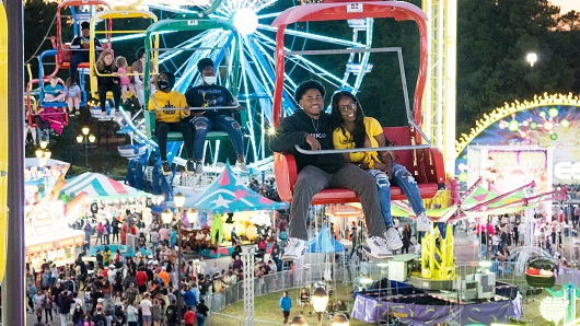 A young couple ride the State Fair Flyer high in the air at night with colorful lights from rides and a Ferris wheel behind them and lots of people milling about on the ground in the distance 