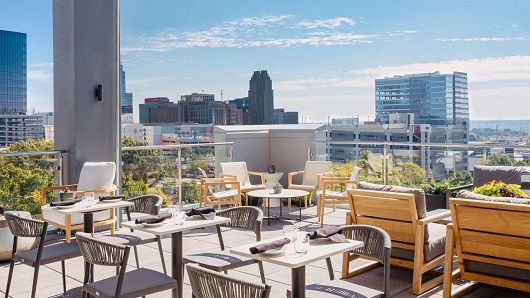Covered patio in daytime with tables and chairs setup for dining; view of Raleigh skyline in distance