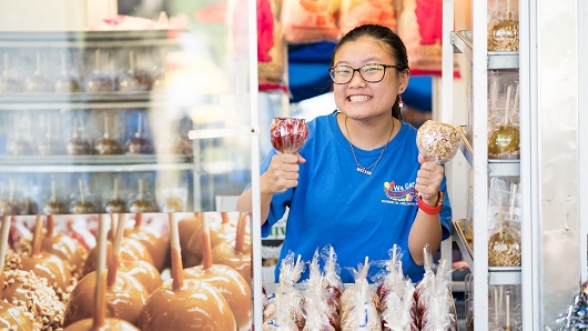 Asian woman in blue shirt and glasses working at food both smiles at the camera while holding up two different styles of candy apples