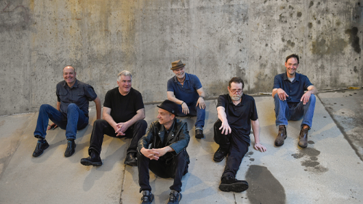 Photo of a six-person rock band sitting in front of a cement wall