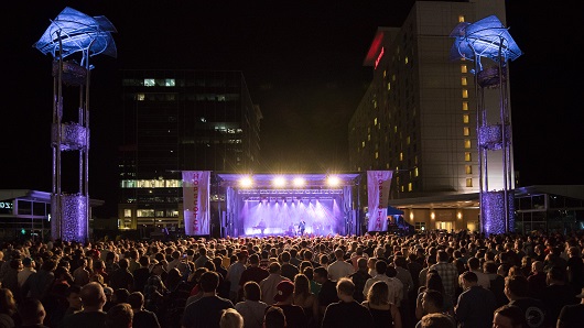 Nighttime scene of large crowd facing music stage outdoors between buildings in downtown Raleigh, with purple and blue lights coming from stage and surrounding light towers