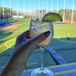 First-person view from a woman holding a cocktail glass up with a lime garnish with a driving range in the background