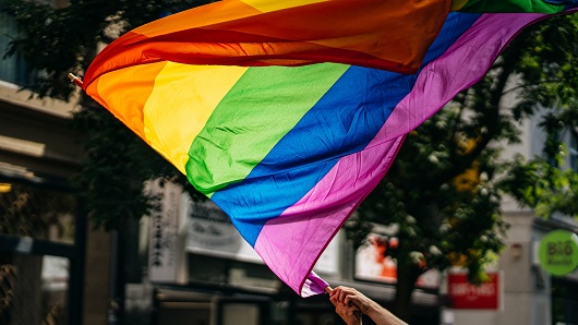Up-close shot of a Pride flag being held in the air on a sunny day within a city setting