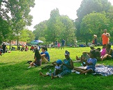 Groups of people of varying sizes enjoy picnics on the lawn at Dix Park, with shade provided by large trees filled with green leaves