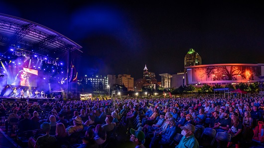 Big crowd fills seats at nighttime show at Red Hat Amphitheater, with stage to camera left and the Raleigh Convention Shimmer Wall glowing in red in the background
