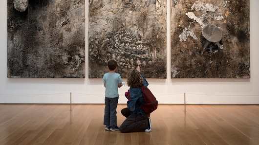 A woman kneels down next to a small child and points at a large piece of art within a gallery that they both are gazing at