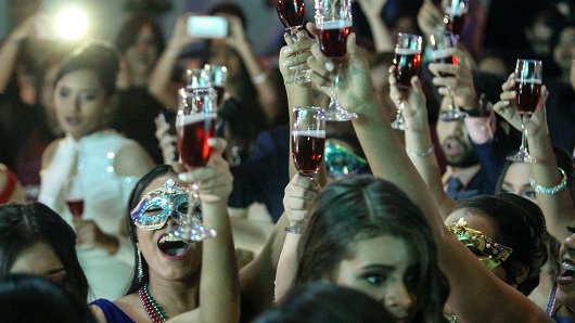 A group of party goers raise glasses of a red-colored drink in the air for a toast; some of the people are wearing masquerade attire