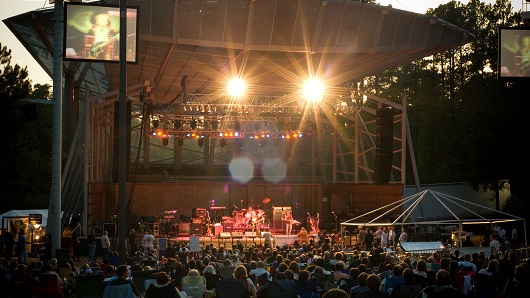 A crowd gathered at Koka Booth Amphitheater in Cary, N.C., with bright lights shining from stage where large band is setting up