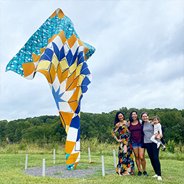 Three women posing next to a giant, kite-looking, colorful outdoor piece of art