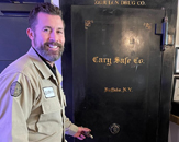 Smiling caucasian man stands in front of vintage safe with hand on handle as he opens; at top of safe reads Zebulon Drug Co., photo is taken at Olde Raleigh Distillery in Zebulon, N.C.