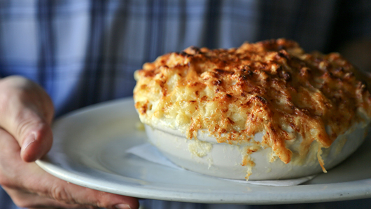 A bowl of mac and cheese sits on a platter held by someone; the mac and cheese has been put under a broiler and has a golden yellow and brown crust on top that spills over the edges