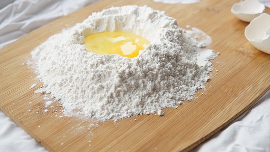 A ring of baking flour sits on a wooded cutting board with eggs cracked into the middle, waiting to be made into handmade pasta