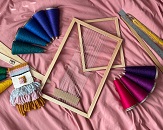 A top-down view of a bundle of tapestry weaving tools sitting on a blush pink fabric; tools include a frame, threads, ruler and more