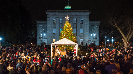 A crowd of people gathered around a large holiday tree in front of the historic North Carolina State Capitol building