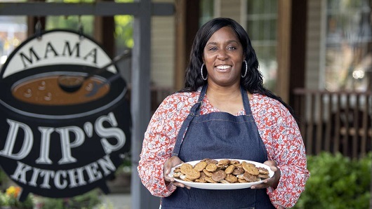 Tonya holding out a plate of cookies with an apron on, in front of a Mama Dip's Kitchen sign