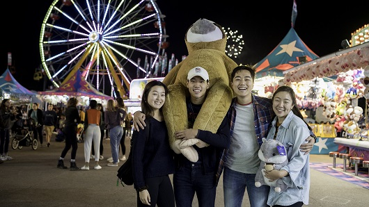A group of four Asian young adults, two men and two women, smile and laugh at camera with the N.C. State Fair glowing at night in the background. One young man holds a giant teddy bear presumably won at a nearby game tent