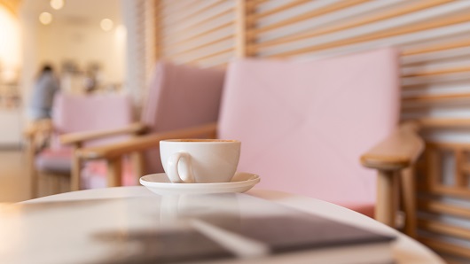 A cup of coffee sits on a table with books, with pink-cushioned chairs in the background and warm light coming from the bar area
