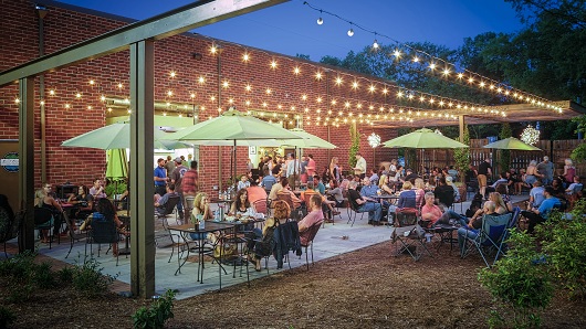 A busy patio at the brewery; tables that have soft green umbrellas are filled with people; string lights are glowing in the dusk light