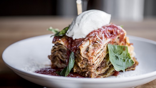 One delicious looking serving of lasagna sits on a white plate, with basil and whipped ricotta on top 