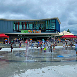 A vibrant celebration with kids playing in a splash pad in front of a new-like, glass-covered building
