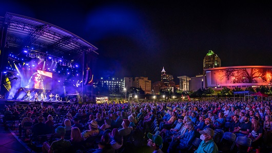 Wide angle shot of huge crowd at Red Hat Amphitheater, with artist playing on stage to left and Raleigh skyline in background and to the right; neon lights shine against nighttime backdrop