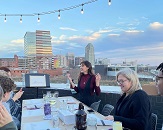 Artist Lauren stands in front of easel and a view of the Raleigh skyline while talking and teaching a table of workshop students how to watercolor an iconic Raleigh scene