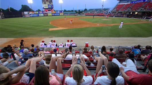 Looking at baseball field from seating along third base; group of fans in foreground with feet up on railing while game is being played at dusk