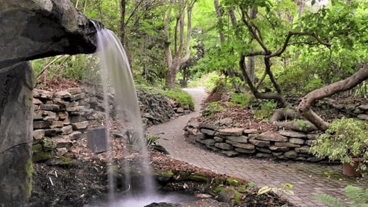 A small waterfall of about 7 feet flows freely in the foreground of an outdoor garden, with a stone walkway winding away from the camera in the background