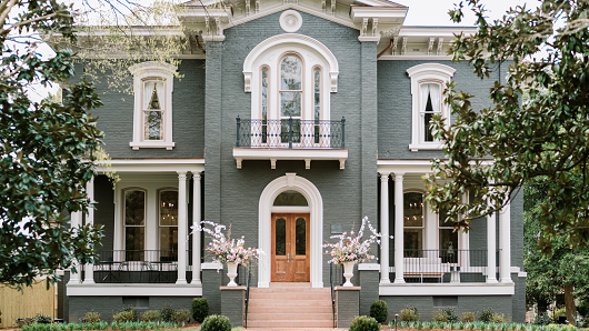 The front view of Heights House Hotel, a historic mansion in a grayish-green brick color with white trim and a front door centered between two white vases filled with white and pink flowers