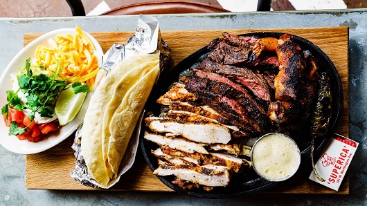 A top-down view of a Tex-Mex platter ready to be made into tacos: grilled chicken, tortillas, cheese, tomatoes, cilantro, lime and more