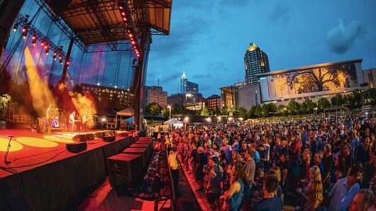 View from side of stage at Red Hat Amphitheater, with large crowd standing and watching band and view of Raleigh skyline at dusk in the background