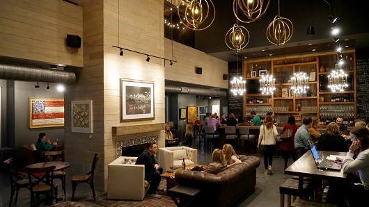 A cozy, dimly-lit room with a wine bar, a fireplace in the center and people lounging on white and brown lounge chairs and couches