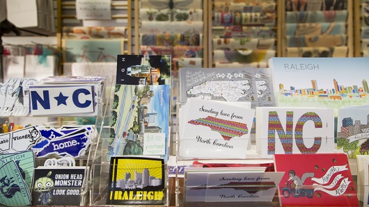A shelf in a retail shop willed with Raleigh and North Carolina-related items, including postcards, stickers, keychains and more