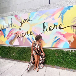 A women in front of a colorful, large outdoor mural that says Wish You Were Here