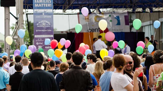 A festival in an urban setting with large crowd of people standing in front of stage with colorful balloons being held by many people